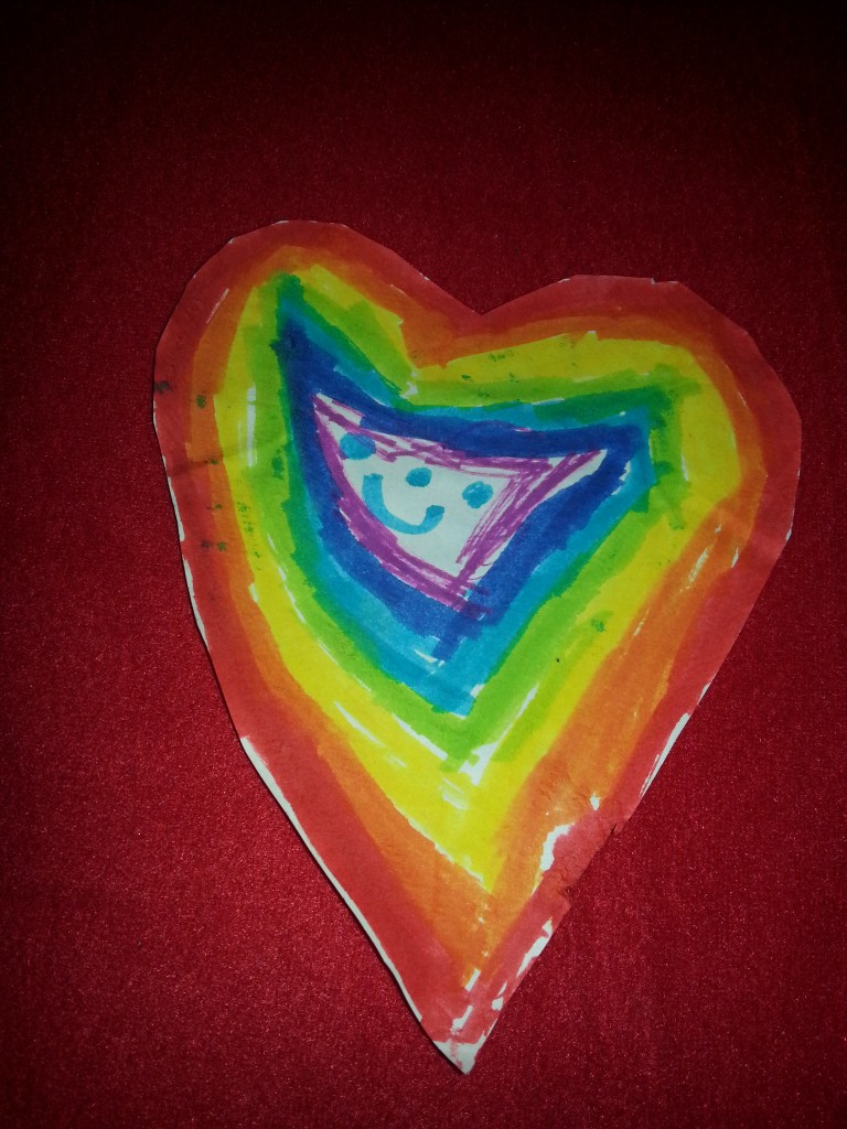 A multi coloured heart with a happy, full of life face in the middle drawn by my young friend Harriet